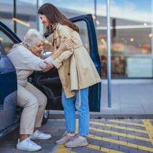 A woman helping an elderly man get into the back of his car.